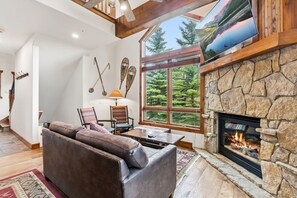A cozy living room with a stone fireplace, a large window with a view of trees, a leather sofa, and a television mounted on the wall. - Breck Basecamp | Deck + Hot Tub + Walk Everywhere (Breckenridge)