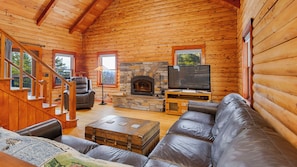 A log cabin interior with a stone fireplace, leather sofa, television, and a staircase. - Cedar & Stone: Keuka Lake Retreat w/ Hot Tub & Golf (Hammondsport)