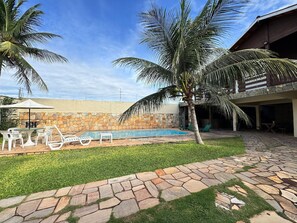 A pool area with stone walls, a grassy lawn, and white lounge chairs. - Casa Imperial (Aquiraz)