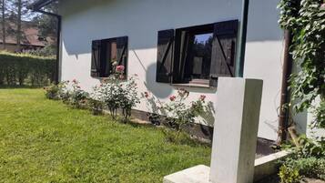 Um edifício branco com duas janelas, cada uma com uma persiana preta. Há um jardim com grama e plantas floridas em frente ao edifício.