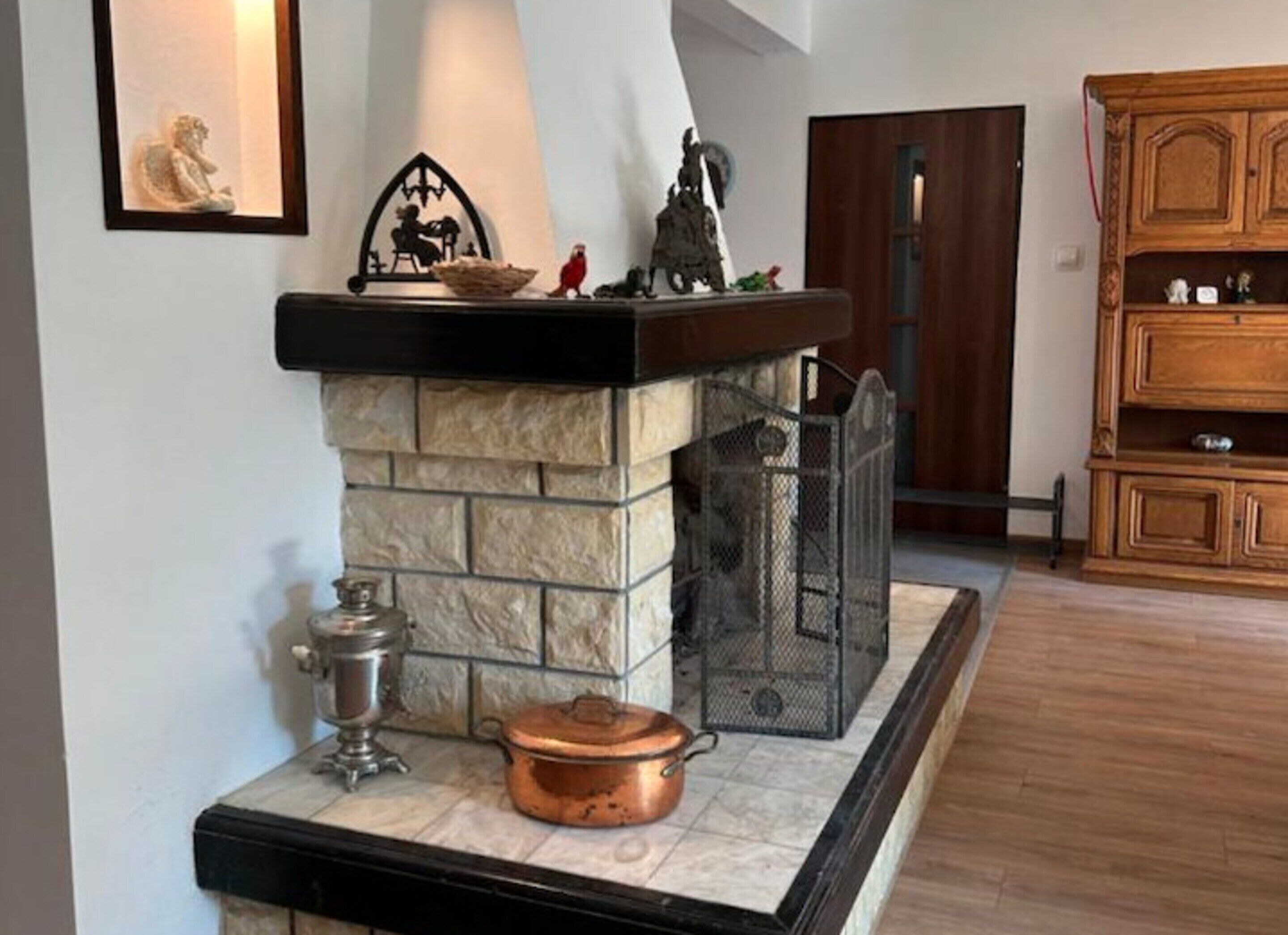 Una chimenea de piedra con una rejilla de metal, una olla de cobre y un jarro de metal sobre un piso de baldosas.