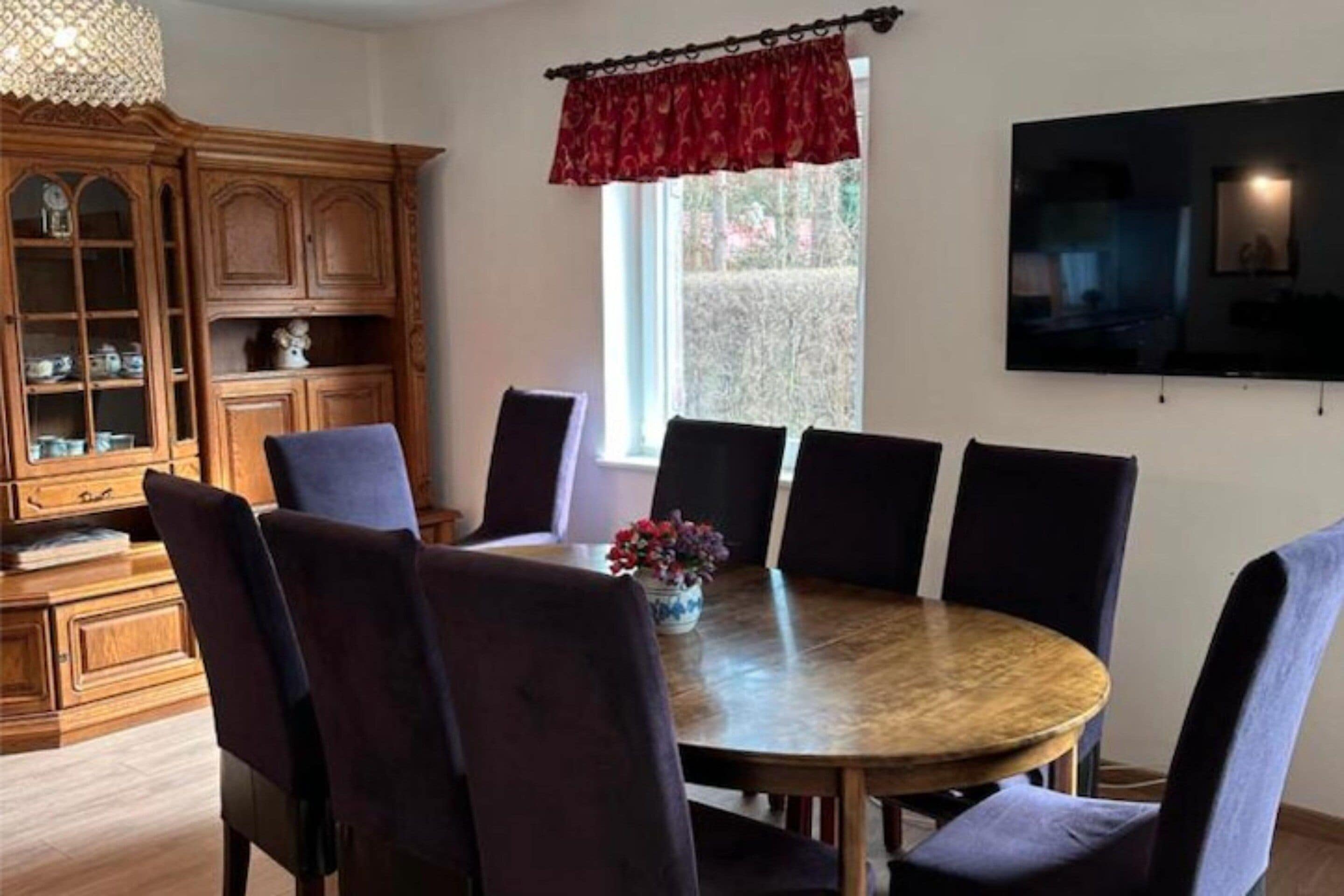 A dining area with a round wooden table surrounded by chairs, a wooden cabinet with glass doors, a window with a red valance, and a flat-screen TV mounted on the wall.