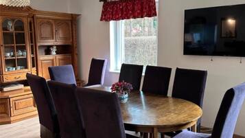 Uma área de refeições com uma mesa redonda de madeira, rodeada por cadeiras, um armário de madeira com portas de vidro, uma janela com cortina vermelha e uma TV de tela plana fixada na parede.