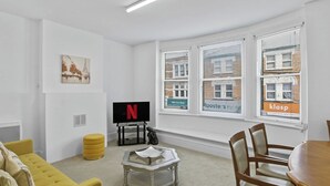 A modern living room with a flat-screen TV, a yellow ottoman, a coffee table, and a dining area with wooden chairs. - Elegant 3BR | Short London Commute | Fast Wi-Fi (Greater London)