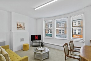 Ein modernes Wohnzimmer mit Flachbildfernseher, einem gelben Hocker, einem Couchtisch und einem Essbereich mit Holzkästen.