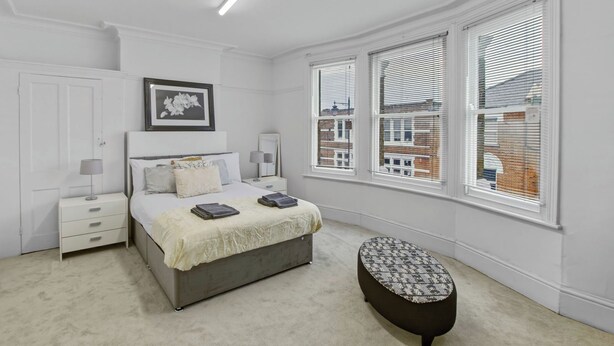 A bedroom with a bed, bedside tables, a bench, and large windows with blinds. - Elegant 3BR | Short London Commute | Fast Wi-Fi (Greater London)
