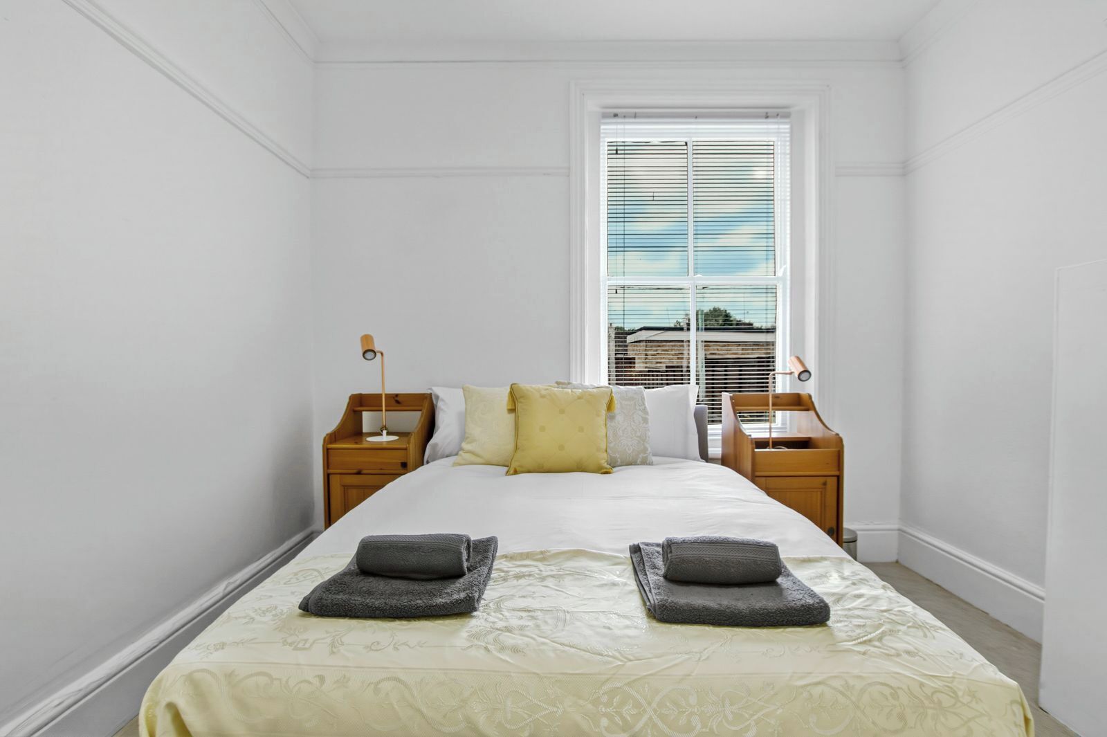 A bedroom with a bed, two nightstands, a window with blinds, and a view of buildings outside.