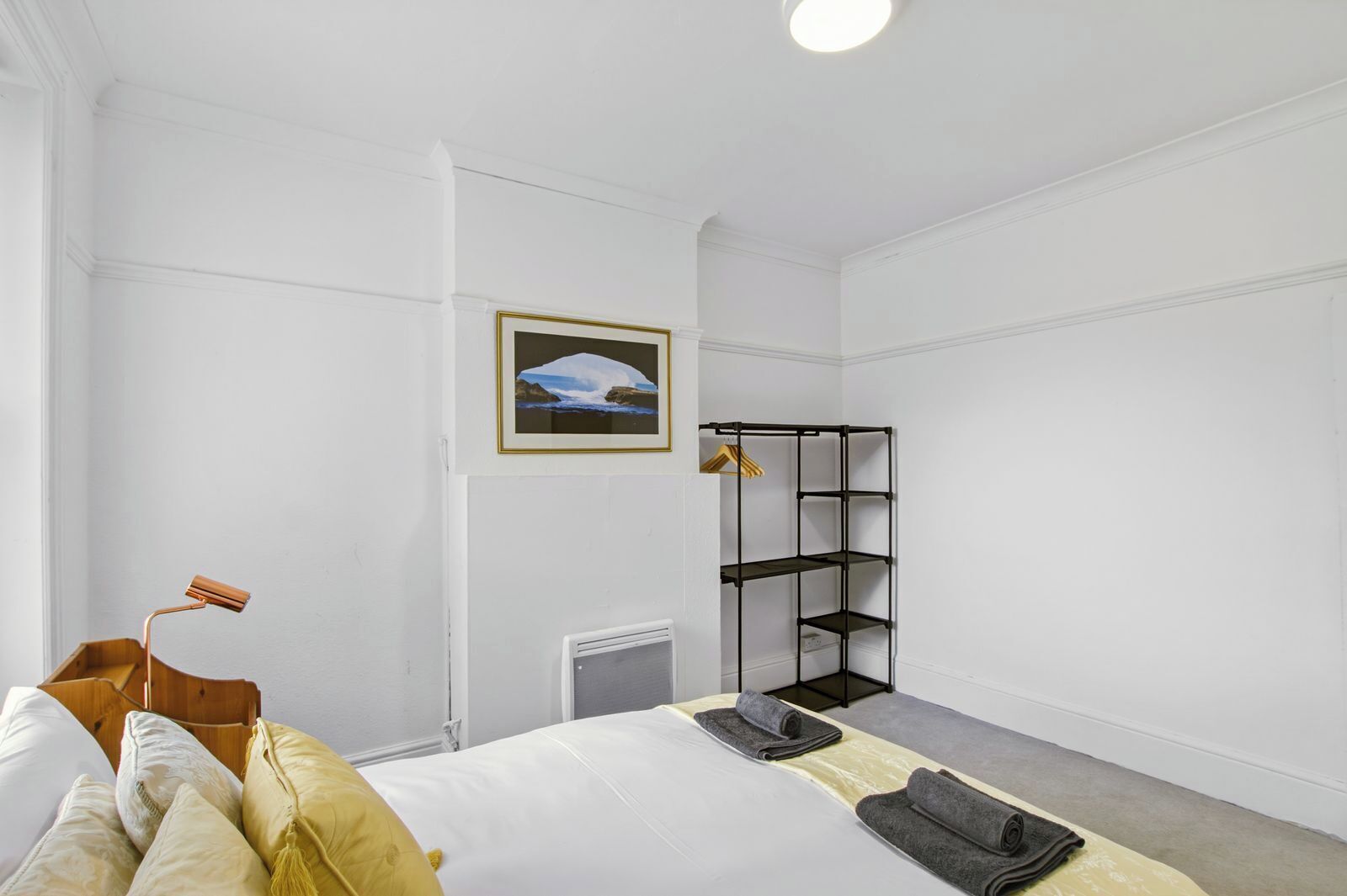Une chambre d’hôtel avec un lit, un radiateur, une étagère en métal noir et un tableau encadré au mur.