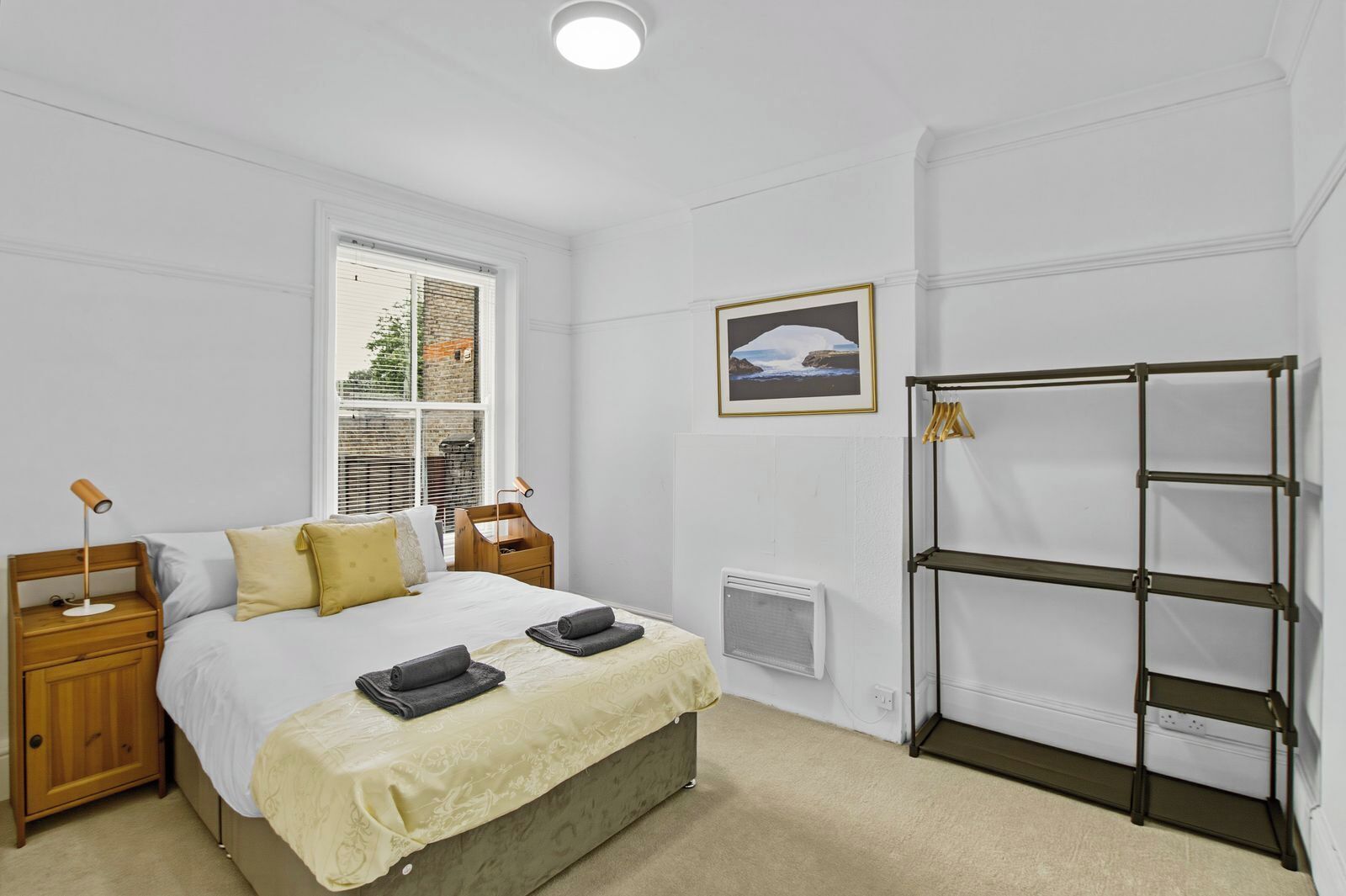 A bedroom with a bed, bedside tables, a window with blinds, a radiator, a wall-mounted shelf, and a framed picture.