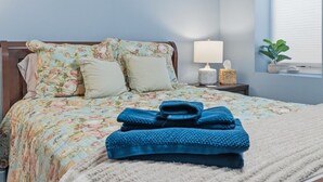 A neatly made bed with floral bedding, a bedside lamp, a small plant, and a folded blue towel on the bed. - Modern Reynoldsburg Retreat Near Columbus (Reynoldsburg)