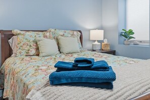 Un lit bien fait, avec une literie à motifs floraux, une lampe de chevet, une petite plante et une serviette bleue pliée posée sur le lit.