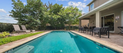 Une piscine rectangulaire à l’eau d’un bleu limpide, entourée d’une terrasse dallée. On y trouve des chaises longues et un espace de détente couvert avec des tables, situé sur le côté droit.