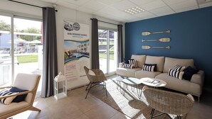 A modern hotel room with a large window, a sofa, a chair, a small table, and a wall with paddleboards.