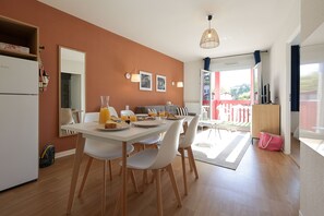A dining area with a white table, chairs, and a refrigerator. There are two framed pictures on the wall, a hanging light fixture, and a view of the outside through a glass door.