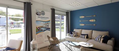 A modern hotel room with a large window, a sofa, a chair, a small table, and a wall with paddleboards.