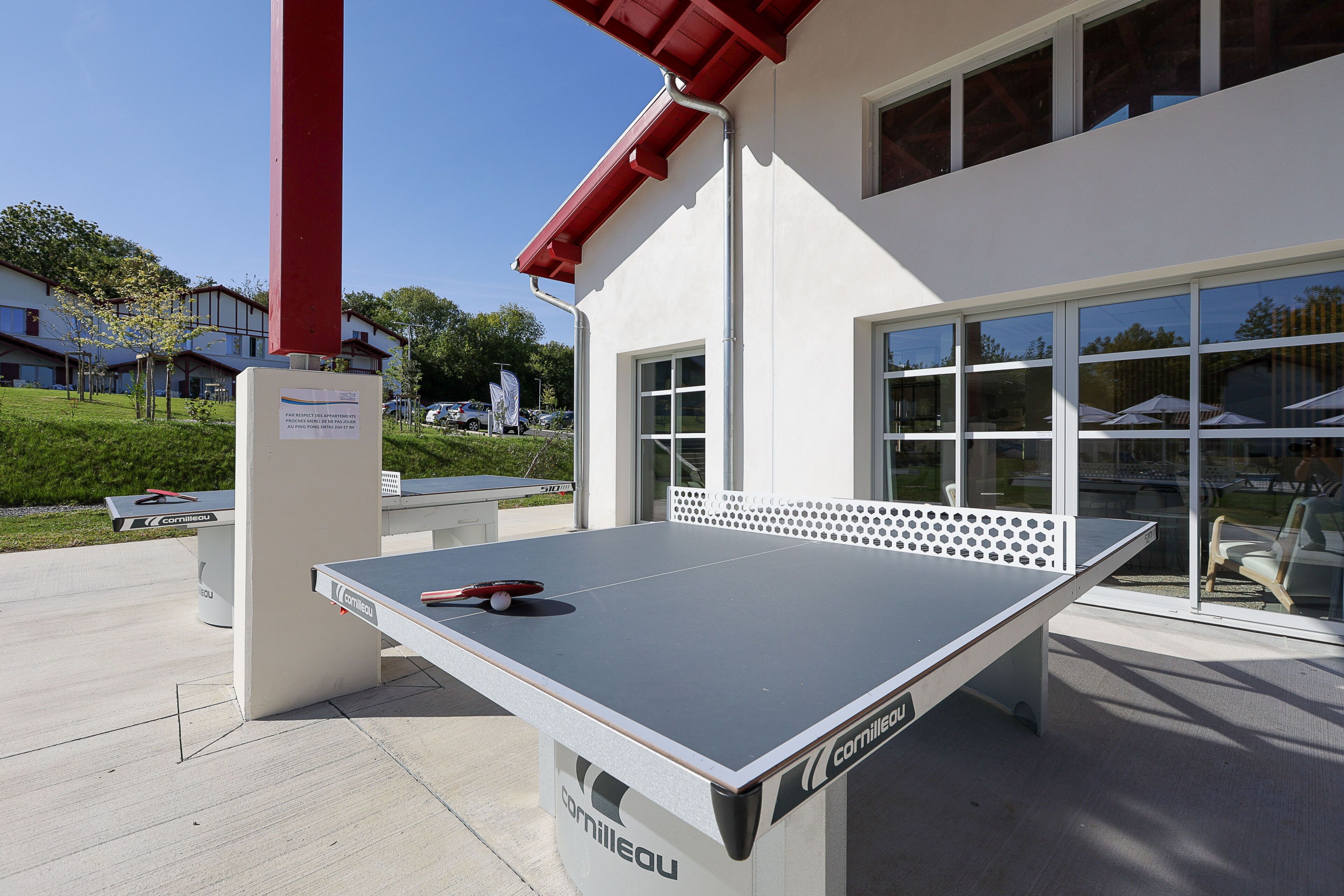 Una mesa de ping pong al aire libre con superficie blanca y detalles rojos, ubicada frente a un edificio con amplios ventanales y tejado rojo.