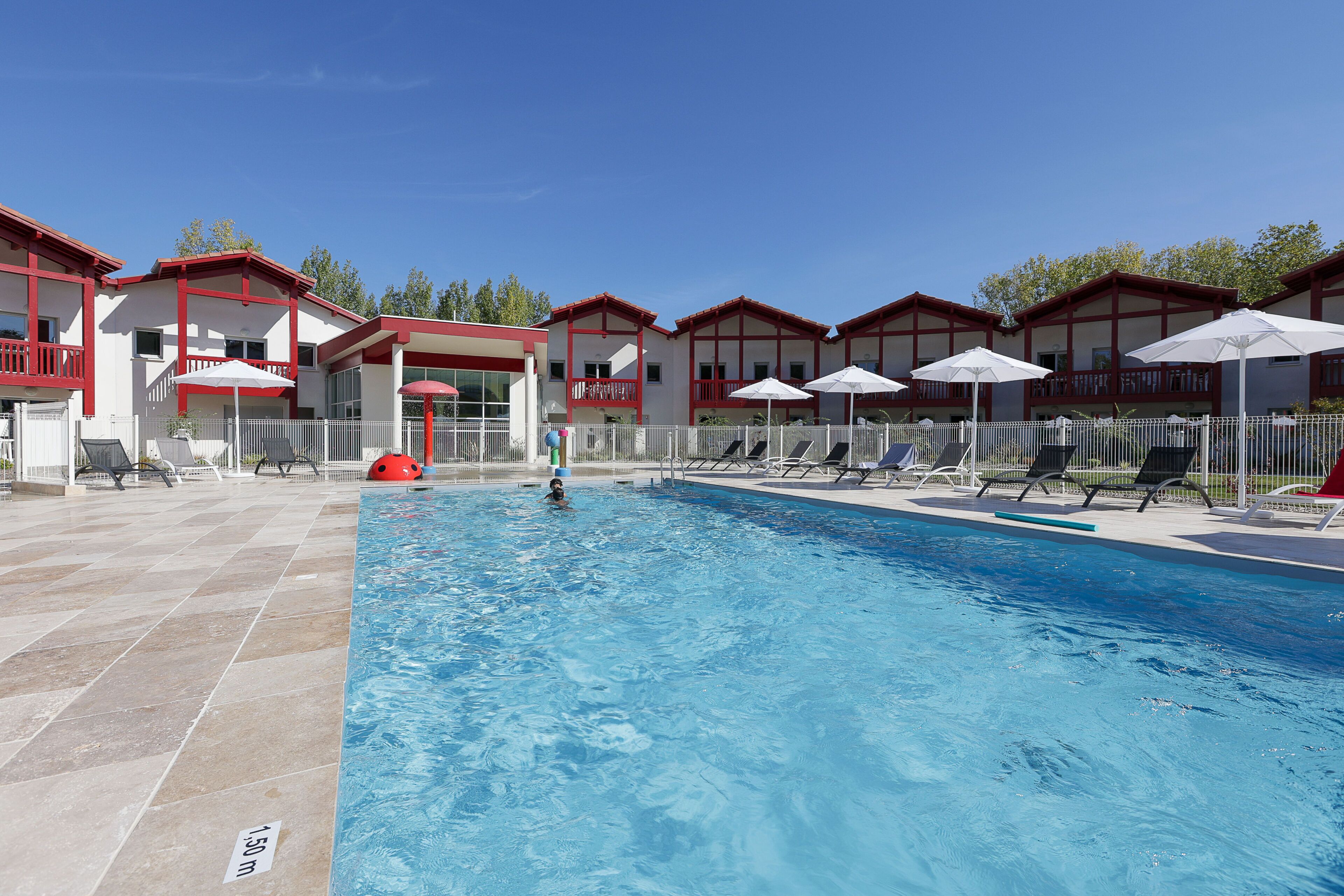 Una piscina con sillones de playa y sombrillas, rodeada de edificios con balcones.