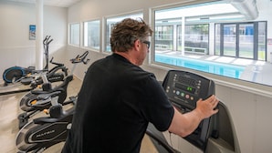 Un hombre usando una cinta de correr en un gimnasio con vista a una piscina y zona de estar al aire libre.