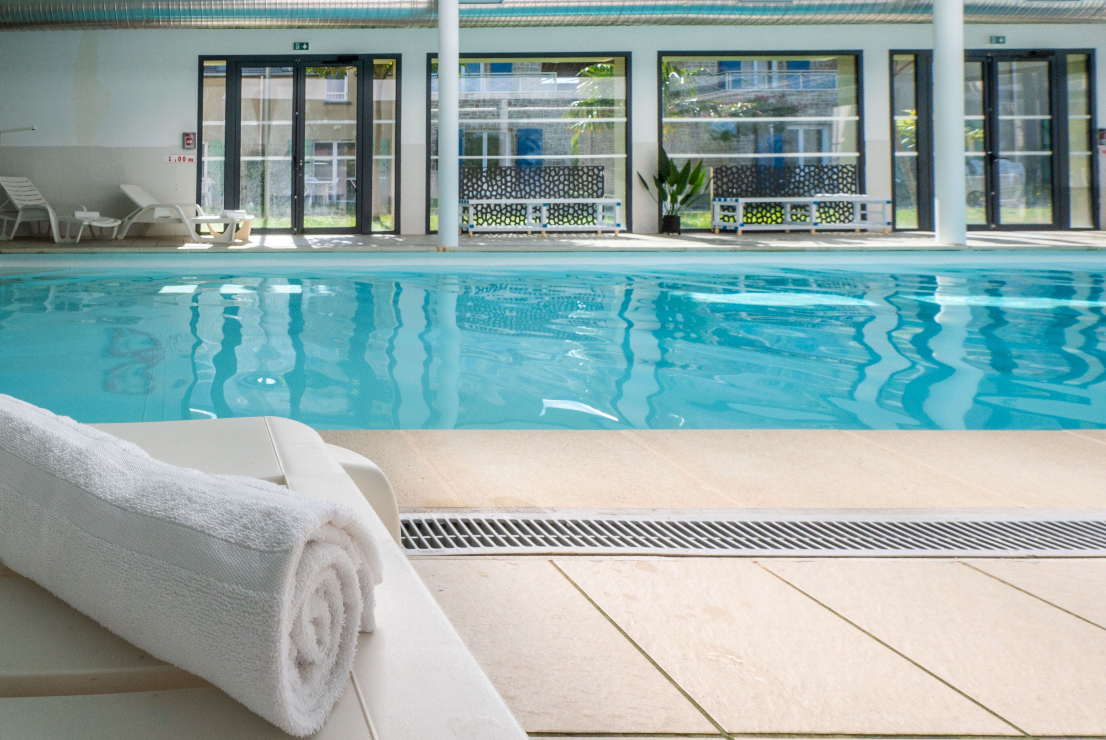 Une piscine avec des chaises longues et des serviettes, située dans un espace intérieur doté de grandes fenêtres offrant une vue sur l’extérieur.