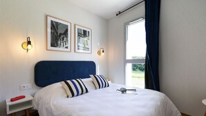 A neatly made bed with white bedding and striped pillows, a bedside table with a red object, and two wall-mounted light fixtures. - T4 spacieux avec accès piscines & wifi inclus (Urrugne)