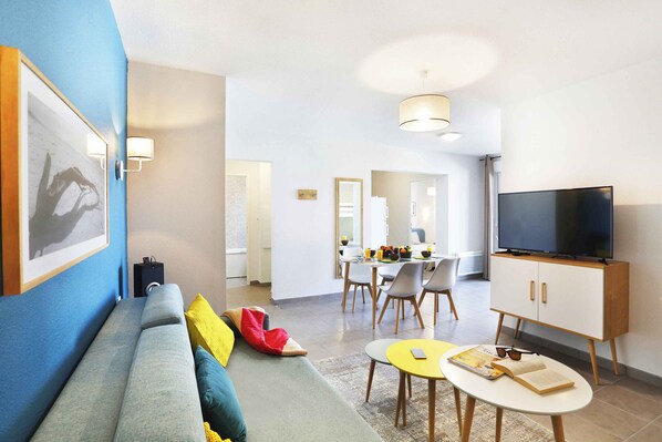 A modern living room with a sofa, a dining area, a TV, and a kitchen in the background.