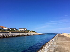 A coastal scene with a concrete walkway along the water, a rocky breakwater, and buildings with red roofs.