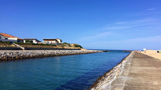 A coastal scene with a concrete walkway along the water, a rocky breakwater, and buildings with red roofs.