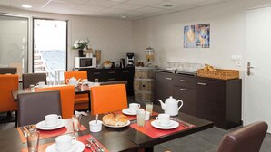 Un coin repas avec une table mise pour le petit-déjeuner, comprenant des croissants, du café et du thé. On y trouve des chaises orange, un four à micro-ondes et une vue sur la mer par une porte vitrée.
