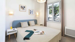A neatly arranged bedroom with a bed, bedside table, lamp, and window with curtains. - À proximité de la plage | Salle fitness | Balcon (Mimizan)