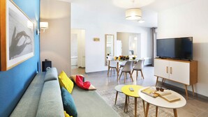A modern living room with a sofa, a dining area, a TV, and a kitchen in the background. - À proximité de la plage | Salle fitness | Balcon (Mimizan)