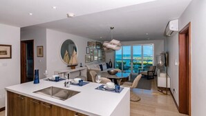 A modern kitchen with a central island, a dining area with a view, and a living space with a large window. - Luxury Apartment with stunning ocean views and pool (St. George)