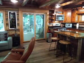 A log cabin interior with a kitchen, dining area, and a large sliding glass door leading to an outdoor patio. - Riverside Serenity: Modern Log Cabin on a Private Trout Stream (Mars Hill)