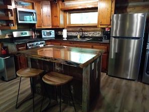 A kitchen with wooden cabinets, a stainless steel refrigerator, a microwave, an oven, and a glass island with stools. - Riverside Serenity: Modern Log Cabin on a Private Trout Stream (Mars Hill)