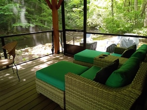 A room with a wooden deck, wicker furniture, and a view of a forested area. - Riverside Serenity: Modern Log Cabin on a Private Trout Stream (Mars Hill)