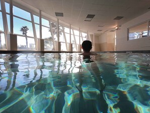 Um homem nada em piscina interior cristalina com grandes janelas e vista para palmeiras.