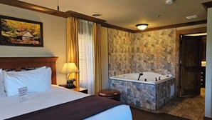 Habitación de hotel con cama, mesita de noche, lámpara, un cuadro, pared de piedra, bañera y puerta.