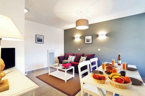 Una sala de estar moderna con sofá, mesa de centro, mesa de comedor con fruta y bebidas, y televisor.