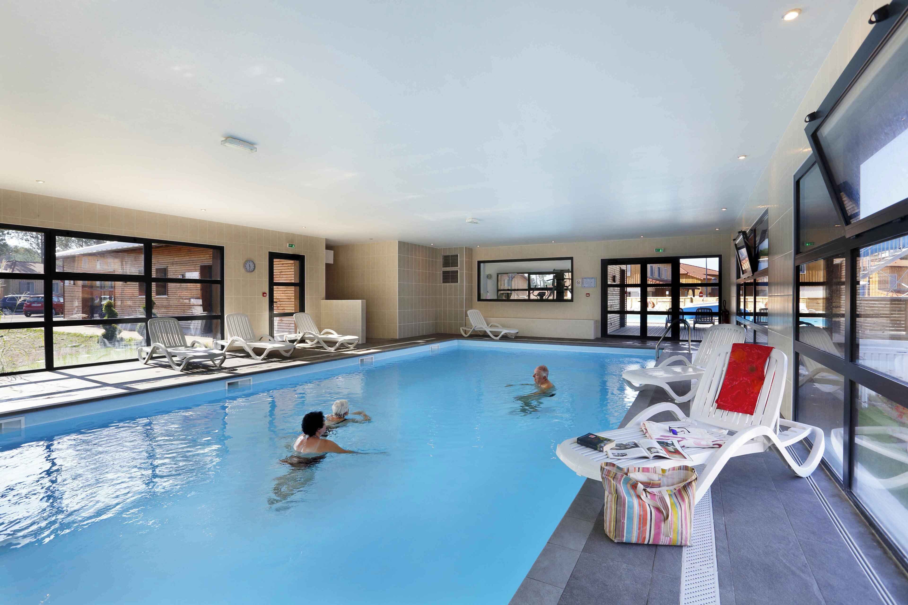 Una piscina interior con paredes de vidrio transparente, sillones de descanso y suelo de baldosas.