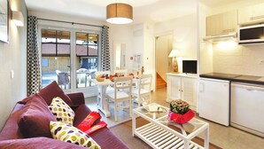 Una sala de estar moderna con sofá, una mesa de comedor y sillas, una zona de cocina con microondas y refrigerador, y ventanas grandes con cortinas.
