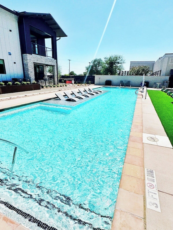 A rectangular swimming pool with a clear blue water surface, surrounded by a paved deck and a grassy area.