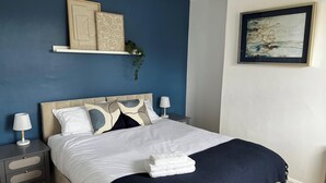 Ein ordentlich bezogenes Bett mit einer weißen und dunkelblauen Bettdecke, ein Nachttisch mit einer Lampe und ein Wandregal mit Dekorationselementen.