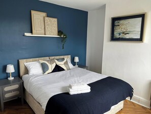 Ein ordentlich bezogenes Bett mit einer weißen und dunkelblauen Bettdecke, ein Nachttisch mit einer Lampe und ein Wandregal mit Dekorationselementen.