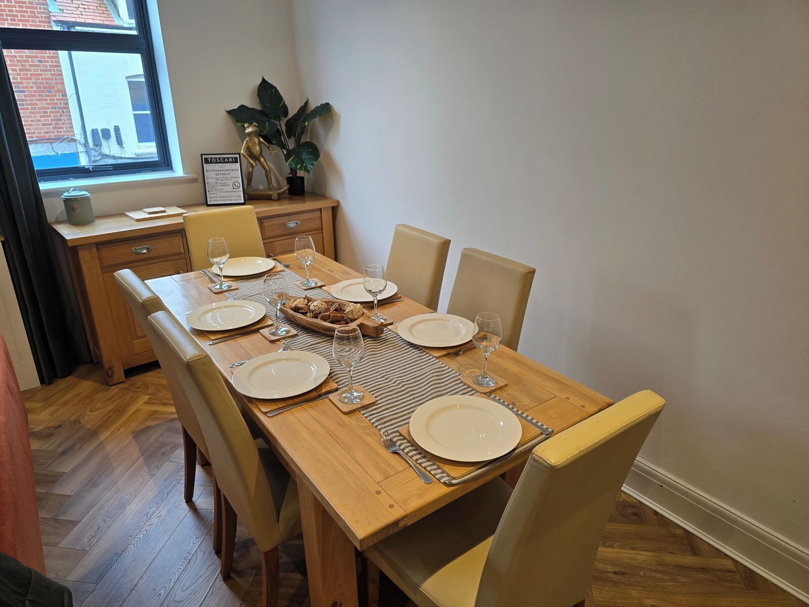 Un coin repas avec une table en bois dressée pour quatre personnes, des chaises, une plante et une vue sur la fenêtre.