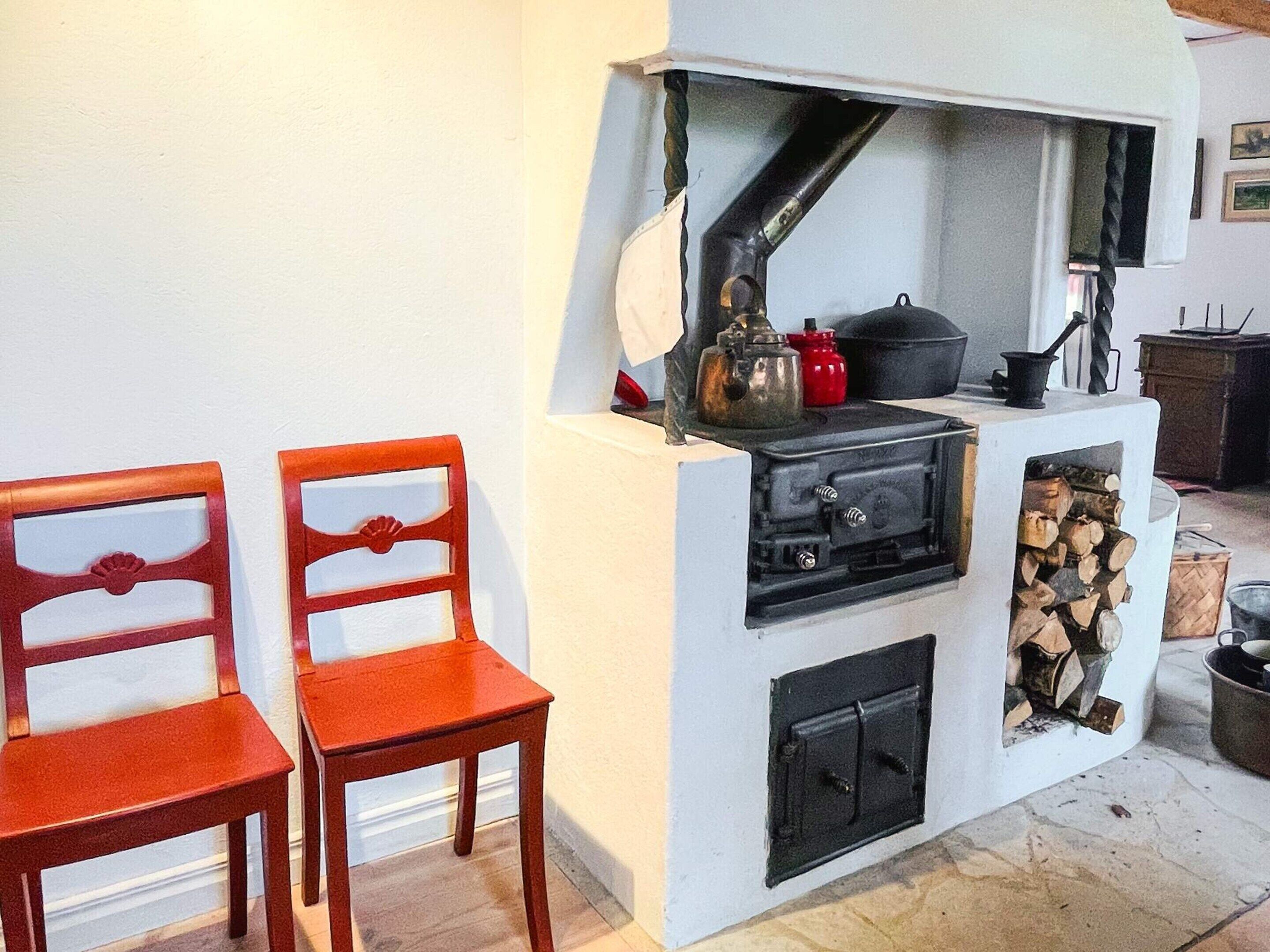 A kitchen with a stove, a pot, a kettle, and a wood storage area. There are two wooden chairs.