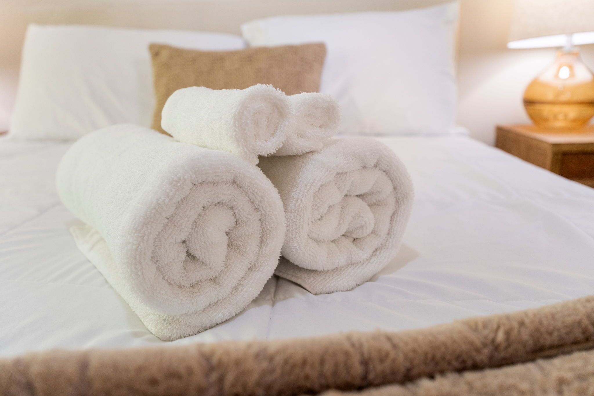 A neatly made bed with rolled white towels, a brown pillow, and a bedside lamp.
