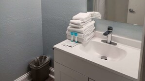 Une salle de bain avec un lavabo blanc, un miroir, une pile de serviettes blanches et une poubelle.