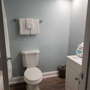A bathroom with a toilet, a towel rack with two towels, a trash can, and a cabinet with toiletries. - Oceanfront Efficiency T1008 (Myrtle Beach)