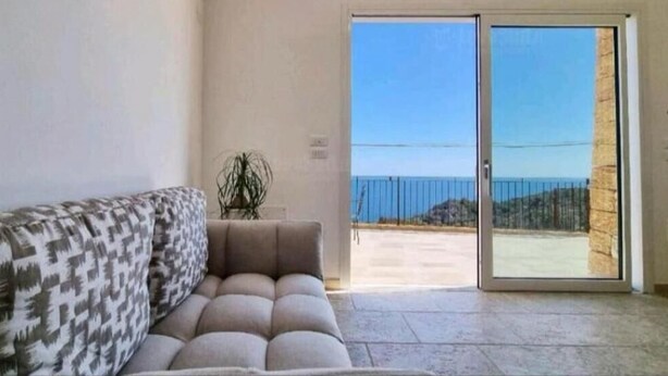Un salón con sofá, planta en maceta y puerta corredera de cristal que da a un balcón con vistas al mar.
