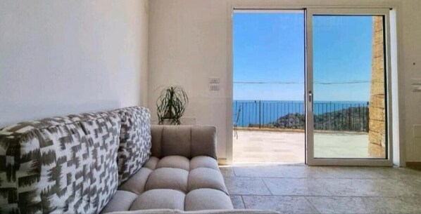Ein Wohnzimmer mit einer Couch, einer Topfpflanze und einer Glastür, die auf einen Balkon mit Meerblick führt.