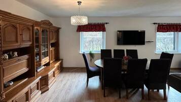 Uma área de jantar com um armário de madeira, um lustre, uma TV na parede e uma janela com vista para árvores.